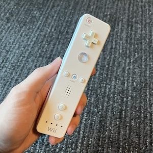 Könnte beinhalten: Eine weiße Wii-Fernbedienung mit einem Steuerkreuz, Tasten und dem Wii-Logo. Die Fernbedienung hat einen Ein-/Ausschalter, eine Home-Taste und Zifferntasten. Die Fernbedienung wird in einer Hand gehalten.