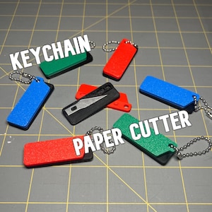 Op de afbeelding: Een verzameling kleurrijke sleutelhangers en papiersnijders. De sleutelhangers zijn rood, blauw en groen, met het woord "KEYCHAIN" in witte tekst. De papiersnijders zijn zwart en rood, met de woorden "PAPER CUTTER" in witte tekst.
