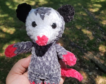 May include: A crocheted plush toy shaped like an opossum. The toy is gray with white and pink accents. It has black ears and a pink nose.