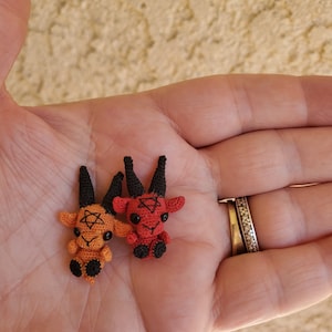 May include: Two small crocheted goat plushies with horns and pentagram designs. One is red and the other is orange.