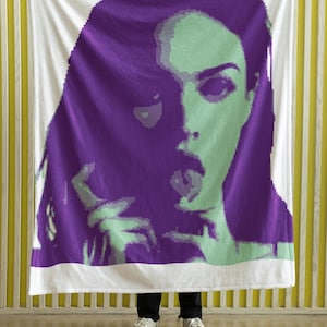 Peut inclure: Une couverture blanche avec une image pixélisée d'un visage de femme en violet et vert. La femme regarde le spectateur et a la bouche ouverte. La couverture est tenue par les mains d'une personne.