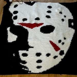 May include: A crocheted blanket featuring a white and black design of a hockey mask. The mask has red accents and is set against a black background.