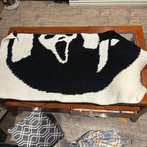 May include: A white and black crocheted blanket with a ghost design. The blanket is laid out on a wooden table.