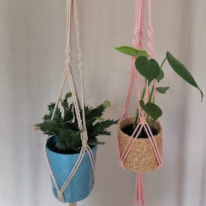 May include: Two macrame plant hangers, one in beige and one in pink, with plants in pots hanging from them.