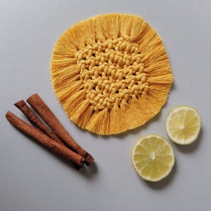 Peut inclure: Un dessous de verre rond en macramé jaune fait à la main, avec un bord à franges. Le dessous de verre est à côté de trois bâtons de cannelle et de deux moitiés de citron sur une surface grise. Le dessous de verre mesure environ 10 cm de diamètre.
