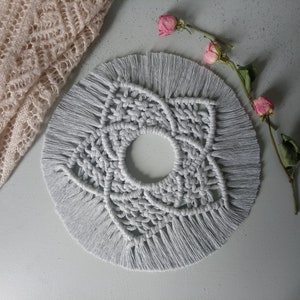 Puede incluir: Un tapiz de macramé gris con un diseño circular y flecos. El diseño es un patrón de estrella con un agujero en el centro.