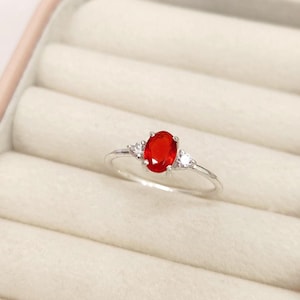 May include: A silver ring featuring a large oval-cut, vibrant red gemstone as the centerpiece, flanked by two smaller, clear, triangular-cut stones. The ring is set against a soft, off-white background, highlighting the jewelry's design.