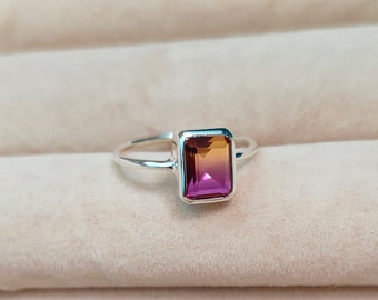 May include: A silver ring featuring a rectangular gemstone with a gradient of colors, transitioning from a deep purple at the base to a golden-orange at the top. The gemstone is set in a rectangular bezel, and the band is a simple, polished silver.