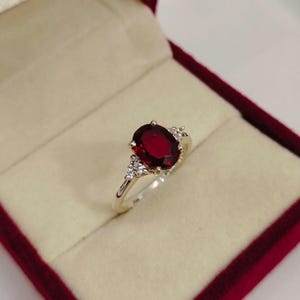 Puede incluir: Un anillo de tono plateado con una gran piedra preciosa de color rojo intenso y corte ovalado como pieza central. La banda está acentuada a ambos lados por un grupo de tres pequeñas piedras claras y brillantes en disposición triangular.
