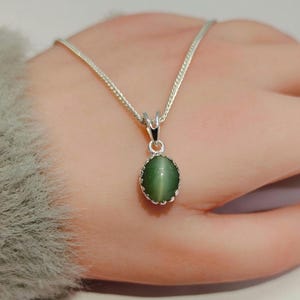 Op de afbeelding: Een zilveren ketting met een ovaalvormige hanger van groene kattenoogsteen. De edelsteen is gezet in een geschulpte zilveren zetting en hangt aan een fijne zilveren ketting. De hanger vertoont een duidelijke chatoyante lichtreflectie.