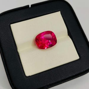 May include: A vivid, cushion-cut, ruby-red gemstone presented in a black jewellery box with a cream-coloured interior. The gemstone's facets reflect light, showcasing its brilliance. The box provides a contrasting backdrop, highlighting the gem's colour and shape.