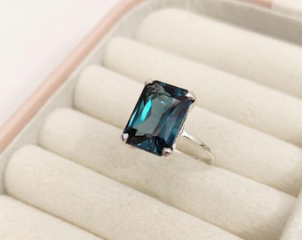 May include: A silver ring with a large, rectangular, teal gemstone. The gemstone is held in place by four prongs. The ring is presented in a white jewellery box.