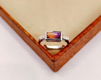May include: A silver ring with a rectangular, multi-colored gemstone. The gemstone transitions from orange to purple. Two smaller, rectangular white stones flank the main stone. The ring is set in a silver band.