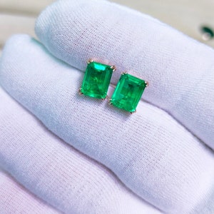 May include: A pair of emerald-cut emerald stud earrings. The vibrant green gemstones are set in a gold-colored metal. The earrings are displayed on a white surface.