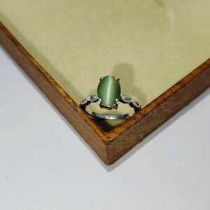 Op de afbeelding: Een zilveren ring met een grote, ovale, olijfgroene edelsteen. De ring heeft een decoratieve band met kleine, heldere accentstenen aan weerszijden van de hoofdsteen. De ring wordt gepresenteerd op een houten oppervlak.