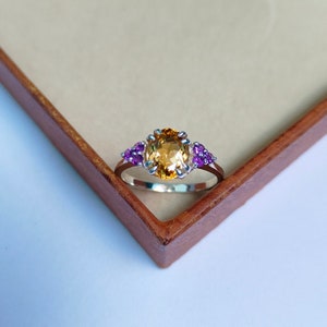 May include: A silver ring featuring a large, oval-shaped, yellow gemstone surrounded by smaller, purple gemstones.