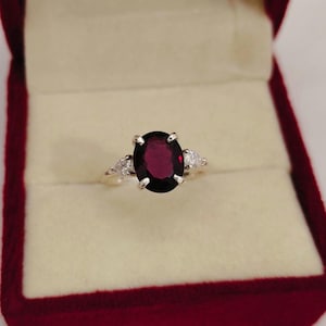 Può includere: Un anello con una grande pietra preziosa rosso scuro dal taglio ovale, montata su una fascia in metallo a quattro punte. La pietra centrale è affiancata su entrambi i lati da una pietra bianca trasparente più piccola a forma di pera.