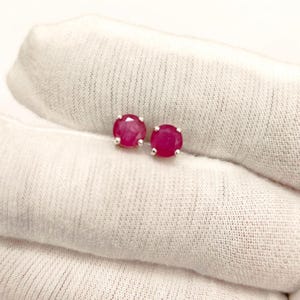 May include: A pair of ruby stud earrings. Each earring features a round, vibrant red gemstone set in a silver-colored metal. The earrings are displayed on a white, textured surface.