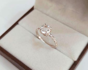 May include: A ring featuring a round, clear, faceted gemstone set in a four-prong setting. The band is crafted from a metallic material and displays an intricate, embossed, vine-like pattern along its outer surface, extending from the setting.