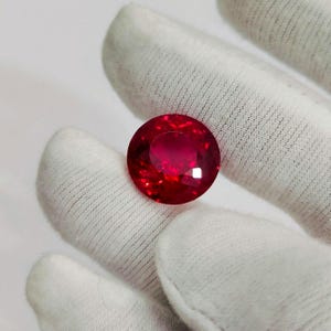 Könnte beinhalten: Ein großer, runder, facettierter Rubin-Edelstein. Der Edelstein hat eine tiefe, leuchtende rote Farbe mit inneren Reflexionen. Der Rubin wird von einer weißen behandschuhten Hand gehalten, die seine Brillanz und Klarheit zeigt. Der Edelstein ist ein Blickfang und suggeriert Luxus und Wert.