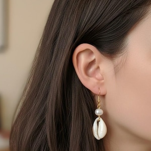 May include: Gold-tone dangle earrings featuring a white seashell and a white pearl. The earrings are suspended from a gold-colored hook. The seashell is a natural, off-white color. The earrings are shown on a person's ear.