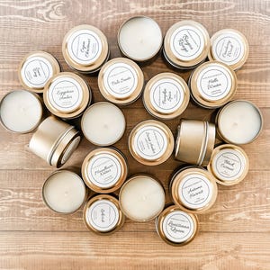 May include: A collection of gold-colored candle tins with various scents, including Egyptian Amber, Palo Santo, Pumpkin Spice, Lakeside, Black Currant, and Louisiana Queen. The candles are arranged in a heart shape on a wooden surface.
