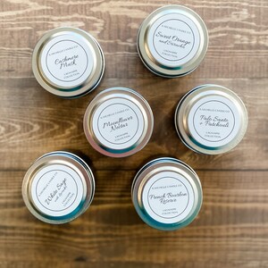 May include: A collection of ten small silver candle tins with white labels. The labels have black text and list the candle scents: White Sage with Rosemary, Cashmere Musk, Sweet Orange and Sriracha, Moonflower Nectar, Palo Santo + Patchouli, and French Bourbon Reserve. All candles are part of the Lagniappe Collection.
