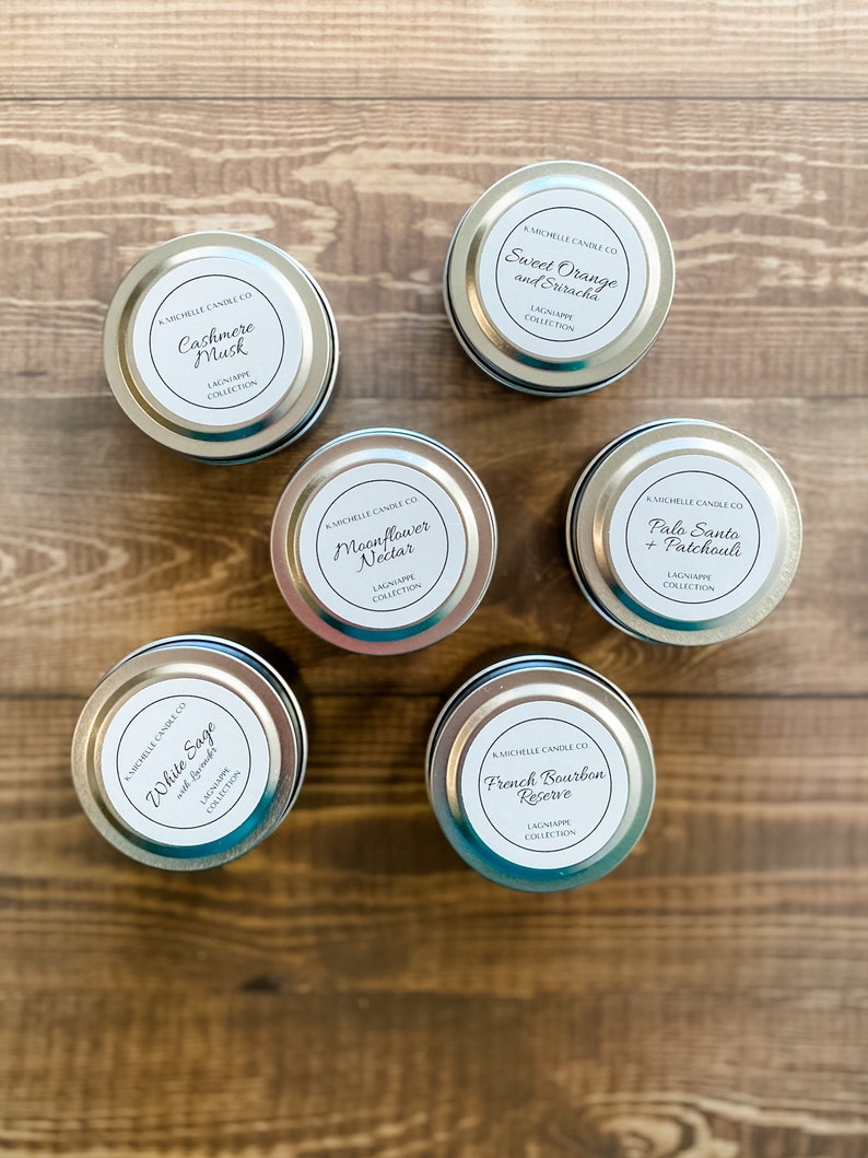 May include: A collection of ten small silver candle tins with white labels. The labels have black text and list the candle scents: White Sage with Rosemary, Cashmere Musk, Sweet Orange and Sriracha, Moonflower Nectar, Palo Santo + Patchouli, and French Bourbon Reserve. All candles are part of the Lagniappe Collection.