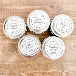 May include: Five silver candle tins with white labels. The labels have black text that reads "KMICHELLE CANDLE CO." and the name of the candle scent. The scents are "Joyeux Noel", "Cajun Christmas", "Fraser Fir", "Hello Winter", and "Cranberry Woods".