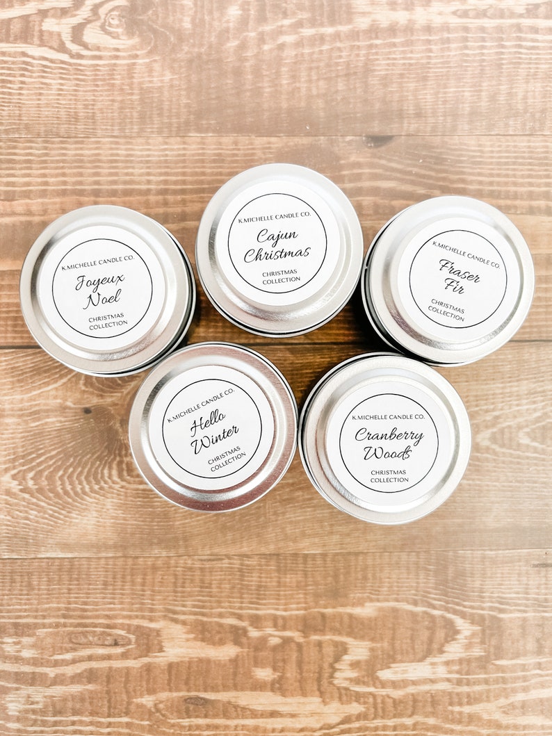 May include: Five silver candle tins with white labels. The labels have black text that reads "KMICHELLE CANDLE CO." and the name of the candle scent. The scents are "Joyeux Noel", "Cajun Christmas", "Fraser Fir", "Hello Winter", and "Cranberry Woods".