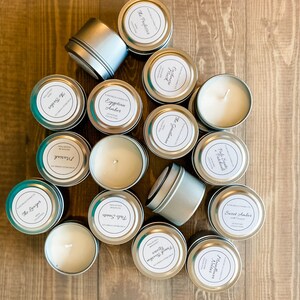 May include: A collection of 15 silver candle tins with white labels. The labels have different names for various scents, including "The Dreamer", "Egyptian Amber", "Sweet Amber", and "Vanilla Cedar".