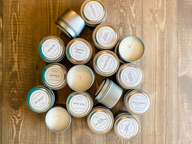 May include: A collection of 15 silver candle tins with white labels. The labels have different names for various scents, including "The Dreamer", "Egyptian Amber", "Sweet Amber", and "Vanilla Cedar".