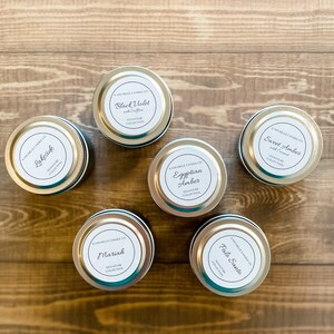 May include: Six silver candle tins with white labels. The labels have black text and list the candle scents: Lakeside, Black Violet with Saffron, Mariah, Egyptian Amber, Palo Santo, and Sweet Amber with Coconut. All candles are part of the Signature Collection.