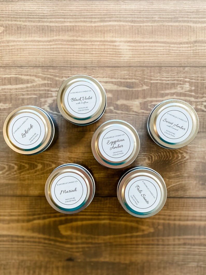 May include: Six silver candle tins with white labels. The labels have black text and list the candle scents: Lakeside, Black Violet with Saffron, Mariah, Egyptian Amber, Palo Santo, and Sweet Amber with Coconut. All candles are part of the Signature Collection.