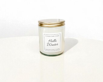 May include: A glass jar candle with a gold lid. The candle has a white label with the text "Hello Winter". The candle is on a white surface.