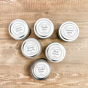 May include: Six silver candle tins with black labels and white text. The labels feature fall-themed scents: Autumn Harvest, Pumpkin Spice, Cinnamon Apple, Granny's Pecan Pie, Black Currant, and Mulled Wine.