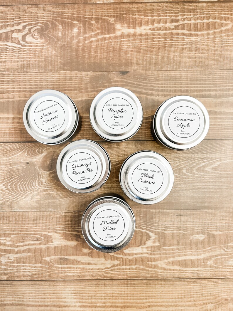 May include: Six silver candle tins with black labels and white text. The labels feature fall-themed scents: Autumn Harvest, Pumpkin Spice, Cinnamon Apple, Granny's Pecan Pie, Black Currant, and Mulled Wine.