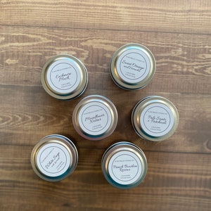 May include: Six silver candle tins with white labels. The labels have the name of the candle scent and the text "Lagniappe Collection". The scents are: White Sage, Cashmere Musk, Moonflower Nectar, Palo Santo + Patchouli, Sweet Orange and Sriracha, and French Bourbon Reserve.