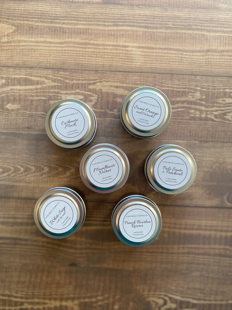 May include: Six silver candle tins with white labels. The labels have the name of the candle scent and the text "Lagniappe Collection". The scents are: White Sage, Cashmere Musk, Moonflower Nectar, Palo Santo + Patchouli, Sweet Orange and Sriracha, and French Bourbon Reserve.