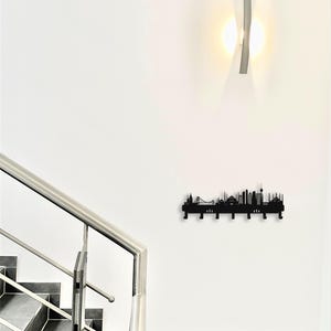 May include: A black metal wall-mounted coat rack featuring a city skyline silhouette design. The rack has multiple hooks for hanging items and is mounted on a white wall. A modern light fixture is above the rack.