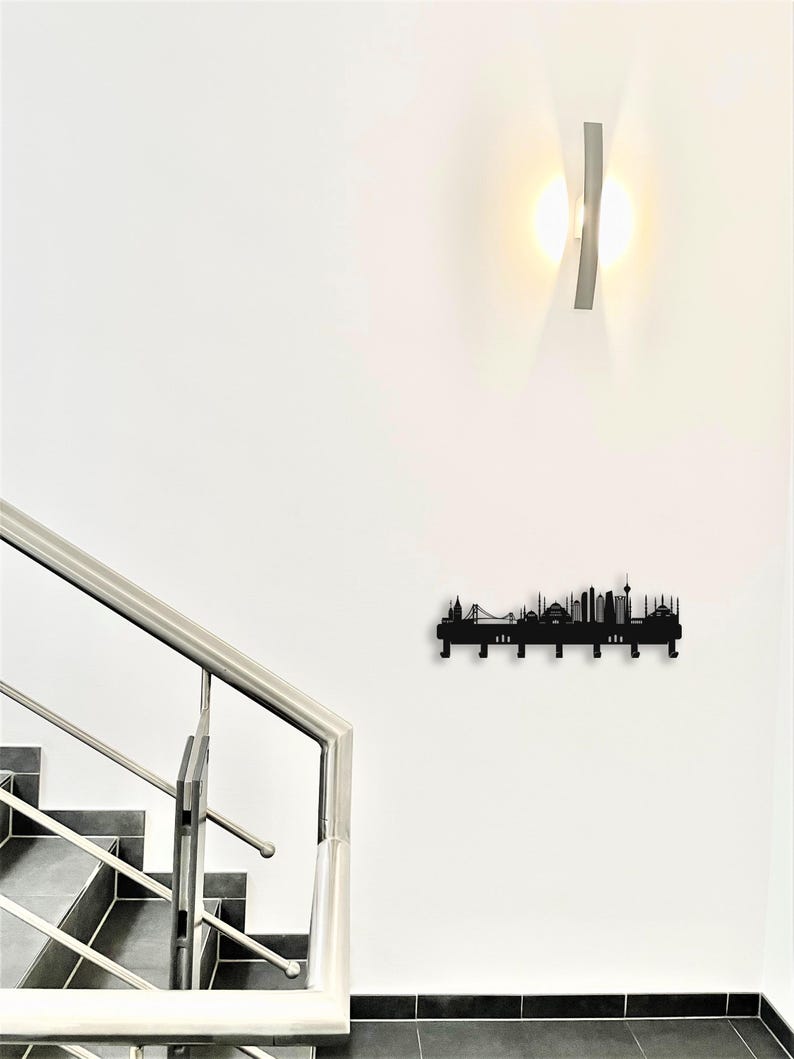 May include: A black metal wall-mounted coat rack featuring a city skyline silhouette design. The rack has multiple hooks for hanging items and is mounted on a white wall. A modern light fixture is above the rack.