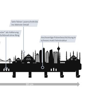 May include: Black metal wall-mounted key holder featuring a skyline silhouette with hooks. The key holder is 12 cm high and 30 cm wide. The design includes architectural details and the text "metallmanufaktur".