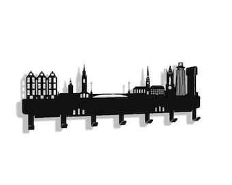 May include: Black metal wall-mounted key holder featuring a city skyline silhouette. The design includes various building shapes, bridges, and spires, with several hooks for keys. The holder is mounted on a white background.