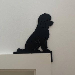 Könnte beinhalten: Eine mattschwarze Silhouette eines sitzenden Pudels, entworfen als Eckdekoration. Der Ausschnitt zeigt die charakteristische lockige Fellstruktur und das Profil eines Pudels und ist so konzipiert, dass er auf der oberen Ecke eines Türrahmens aufliegt.