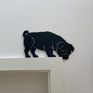 Könnte beinhalten: Eine mattschwarze Silhouette eines Mopses, die als dekorativer Aufsatz für Wände oder Türen gestaltet ist. Die flache, ausgeschnittene Figur zeigt den typischen geringelten Schwanz, das faltige Gesicht und den stämmigen Körperbau eines Mopses.
