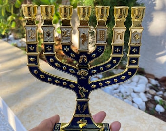 May include: A gold menorah with blue enamel and white accents. The menorah has seven branches and is decorated with Hebrew symbols. The base of the menorah is inscribed with the word "Jerusalem".