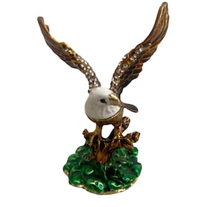 May include: A decorative figurine of an eagle with outstretched wings. The eagle has a white head, brown wings with rhinestone accents, and gold-colored talons. It is perched on a green base.