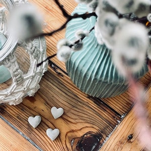 Könnte beinhalten: Drei kleine weiße herzförmige Dekorationen auf einer Holzoberfläche mit einer hellblauen Vase und einem weißen Kranz im Hintergrund.