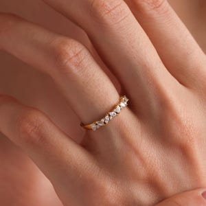 May include: A delicate gold band ring with a row of small, clear gemstones. The ring is worn on the finger of a person with manicured nails.