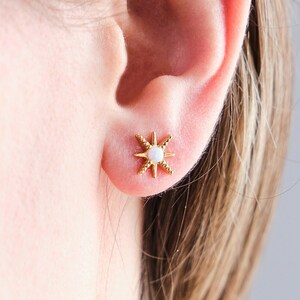 May include: Gold star-shaped earrings with a white opal center. The earrings are shown on a person's ear.